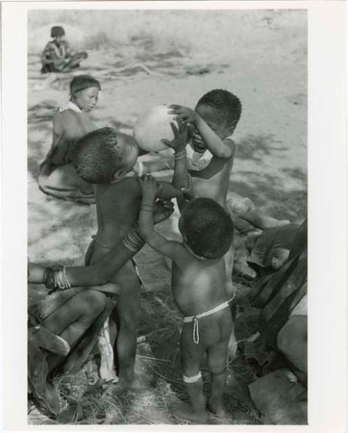 [No folder title]: Debe holding Bau's arms as she drinks from an ostrich eggshell held by /Gaishay while an unidentified, young woman steadies them; unidentified women sit in the background (print is a cropped image)