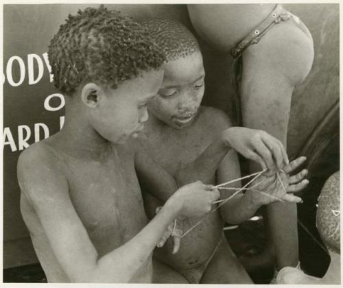 [No folder title]: Two boys making a "cat's cradle" in front of the expedition truck (print is a cropped image)