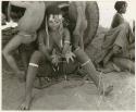 [No folder title]: //Kushay ("/Qui Navel's" wife) making a "cat's cradle" as she sits in front of the expedition truck's tire with unidentified children around her; Di!ai is sitting to the right (print is a cropped image)