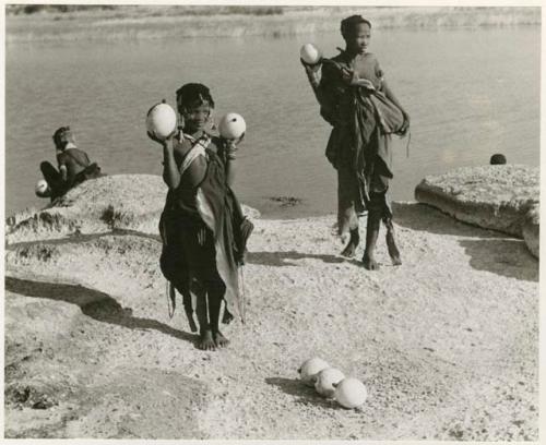 [No folder title]: Two women standing beside Nama Pan holding ostrich egg shells with unidentified people in the background (print is a cropped image)