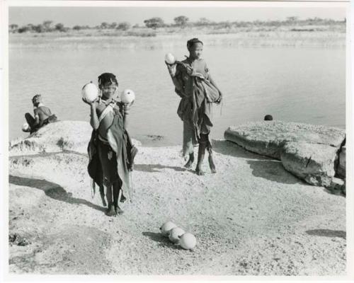 [No folder title]: Two women standing beside Nama Pan holding ostrich egg shells with unidentified people in the background (print is a cropped image)