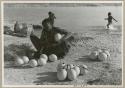 [No folder title]: Khuan//a (Gau's wife) sitting with ostrich egg shells and children playing in the water behind her (print is a cropped image)