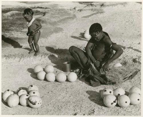 [No folder title]: Khuan//a (Gau's wife) placing ostrich egg shells wrapped in grass in her kaross and her child standing near her (print is a cropped image)