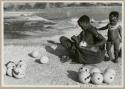 [No folder title]: Khuan//a (Gau's wife) placing ostrich egg shells wrapped in grass in her kaross with her child standing beside her (print is a cropped image)