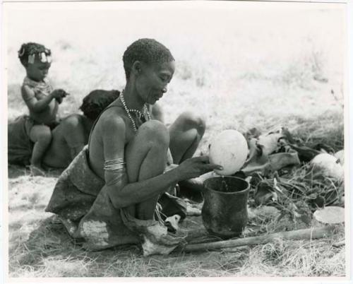 [No folder title]: /Naoka, first wife of "Gao Medicine," emptying an ostrich egg into a pot, with child sitting on somebody's back behind her (print is a cropped image)