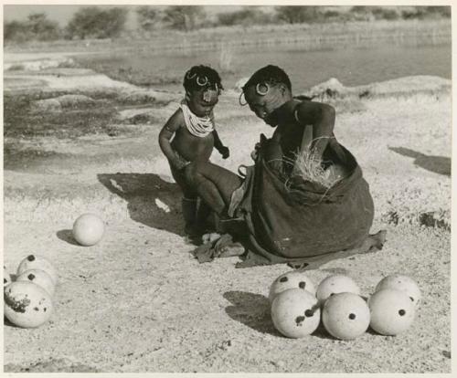 [No folder title]: Khuan//a (Gau's wife) placing ostrich egg shells wrapped in grass in her kaross with her child standing beside her (print is a cropped image)