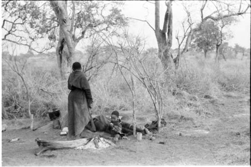 "/Qui Navel" and his son, "Little ≠Gao," lying down beside a fire in the skerm, with //Kushay standing