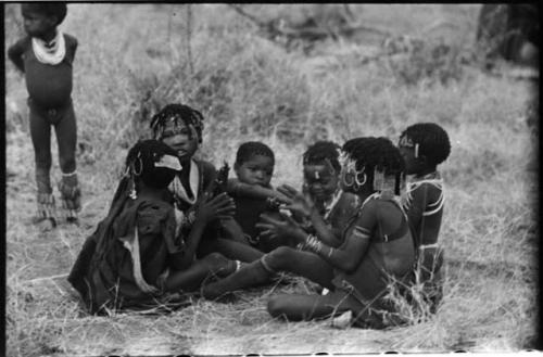 Girls sitting in close circle, clapping and singing; a little boy standing in the background