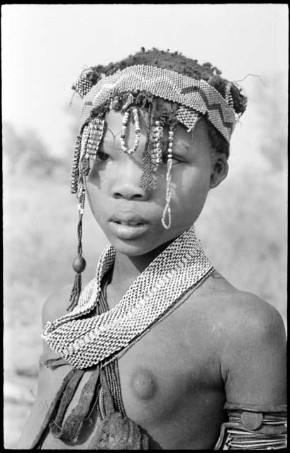 Portrait of N≠isa wearing ostrich eggshell beads around her neck, headband of Western beads on her head, and a long band made of a tsi nut and beads hanging on the right side of her face