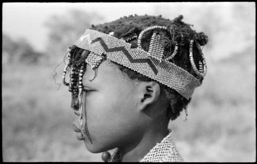 Close-up profile of N≠isa wearing ostrich eggshell beads around her neck, headband of European beads on her head, and a long band made of a tsi nut and beads hanging on the right side of her face