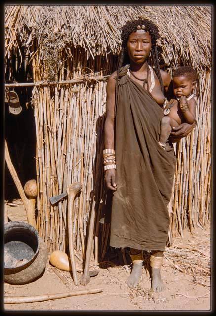 Woman standing and holding a child in front of a hut