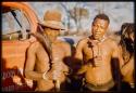 Two men standing in front of the expedition truck, one holding a rattle and the other holding an animal tail