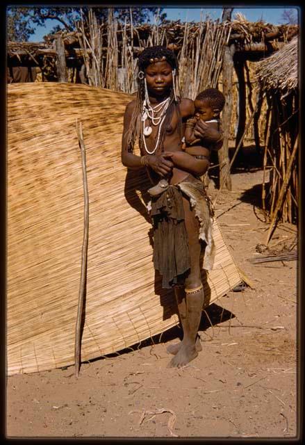 Girl holding a baby, standing in front of a hut