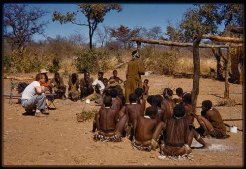 Medicine man dancing, with a group of women sitting and clapping and Nicholas England squatting near them