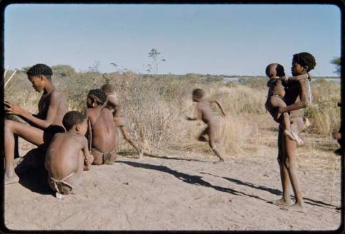 Children, Fathers and sons: Boy carrying a baby up a termite mound, with other boys playing and "≠Gao Lame" sitting (left)