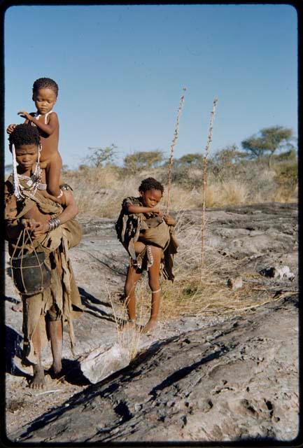 Children, Socialization--hunting, carrying: Woman with two children near a waterhole, carrying one of them on her shoulder and an Ovambo pot in a sling