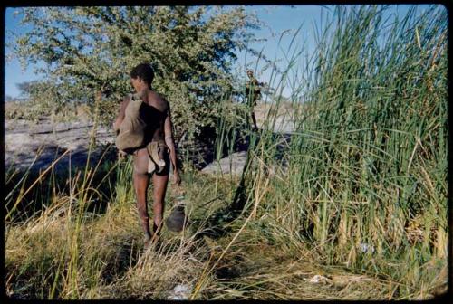 Clothes: Man wearing a breechclout, carrying a bag and an Ovambo pot, approaching a waterhole, view from behind