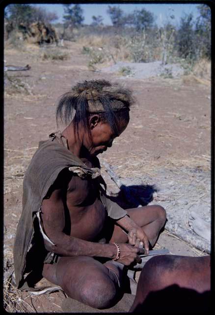 Clothes: "Old !Naishi" wearing a badger cap, sitting with his feet tucked up under him, filing an arrowpoint with a file given to him by the expedition