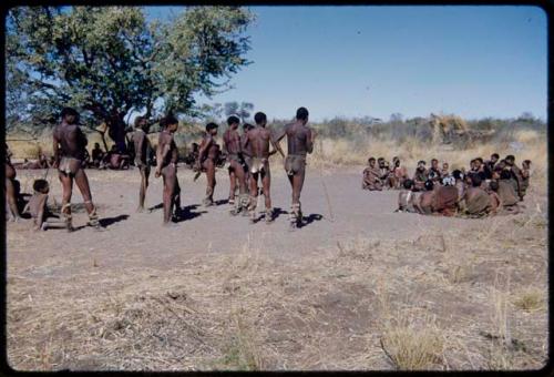 Dance: Men standing, waiting to begin a daytime dance, with women sitting in a circle near them