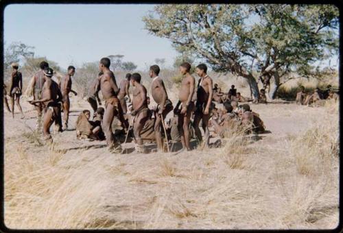 Dance: Men dancing clockwise around a circle of women sitting at a daytime dance, including Gau and ≠Toma (!Naishi's son), with !Naishi walking away