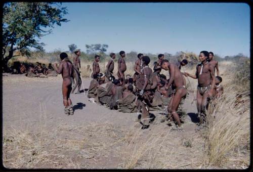 Dance: Men dancing clockwise around a circle of women sitting at a daytime dance, including !Naishi and Gau