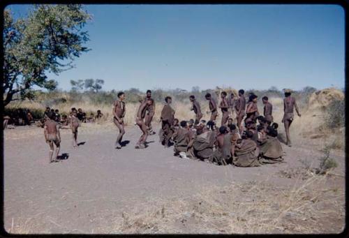 Dance: /Gam leads the line of men dancing counter clockwise through a circle of women sitting at a daytime dance (the primary role of the women is to sit in a circle to clap and sing, providing music for the dancing men)