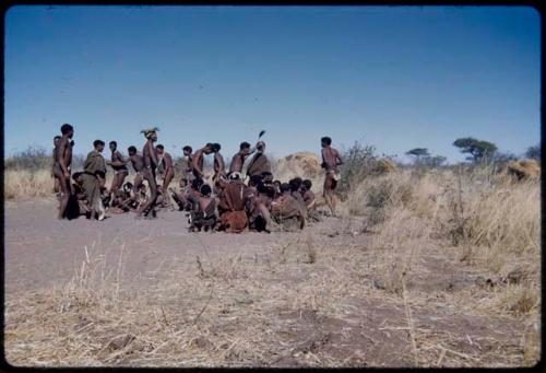 Dance: /Gam leading the men dancing through a circle of women at a daytime dance, with the man behind her holding a wildebeest tail (which men often carry in a dance) over her head (the primary role of the women is to sit in a circle to clap and sing, providing music for the dancing men)