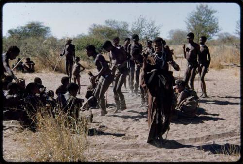 Dance: Men dancing clockwise through a circle of women at a daytime dance, with /Gam about to sit down (the primary role of the women is to sit in a circle to clap and sing, providing music for the dancing men)