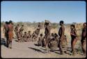 Dance: Line of men dancing counter-clockwise around the circle of women sitting at a daytime dance, close-up