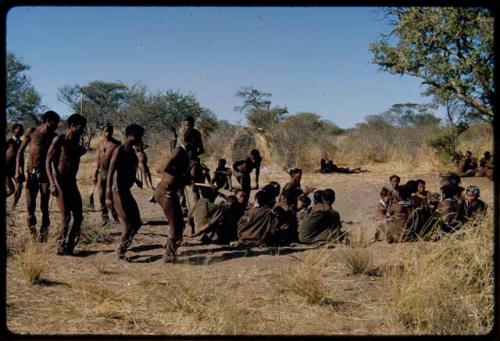 Dance: Line of men dancing counter-clockwise around a circle of women sitting at a daytime dance, close-up, with a man leaning over as though to perform a ritual curing