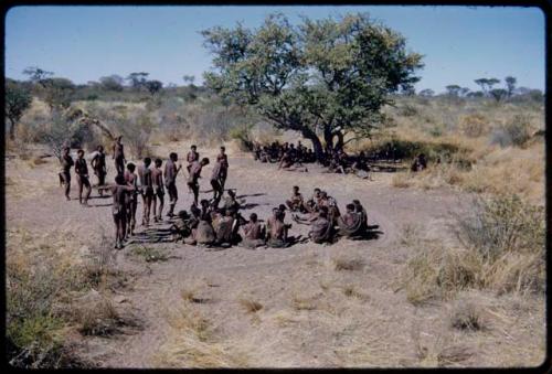 Dance: Men dancing clockwise around the circle of women sitting at a daytime dance, with some men standing, waiting to enter the dance, view from the expedition truck with a wide-angle lens