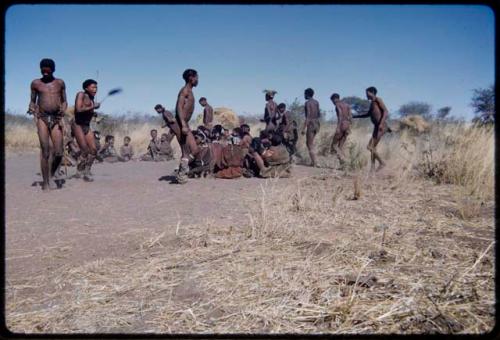 Dance: Line of men dancing counter-clockwise around a group of women sitting at a daytime dance, with /Ti!kay holding "Crooked /Qui's" arms, "Old Di//khao" dancing behind !Naishi and //Ao dancing strongly