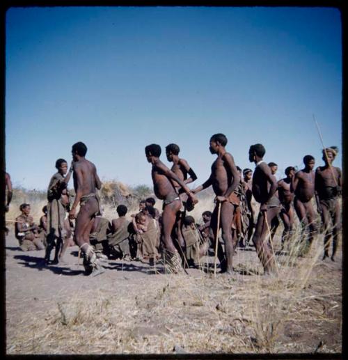 Dance: Line of men dancing clockwise around a group of women sitting at a daytime dance, with "/Gao Music" being held by "Crooked /Qui" and ≠Toma (son of !Naishi) (half of stereo 2001.29.5752)