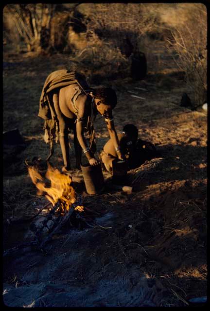 Food, Cooking: Di!ai (second wife of "Gao Medicine") standing and leaning over next to her fire, with her son, /Gaishay, sitting near her