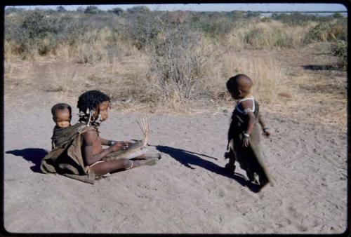 Children, Dancing: N!ai sitting with !Ungka Norna tied to her back, playing the //guashi for /Gaishay (son of "Gao Medicine" and /Naoka) to dance