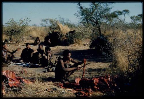 Food, Meat: "Gao Medicine" cutting meat into strips, with his family sitting behind him, including //Khuga (his daughter) in the middle