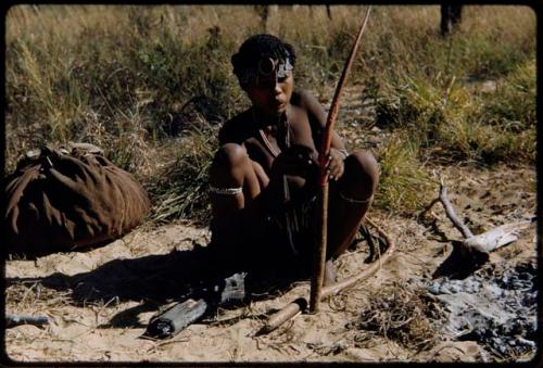 Fiber Root: //Kushay (wife of /Qui) squatting, holding a long orange root from which cord is made, with a kaross filled with something on the ground behind her