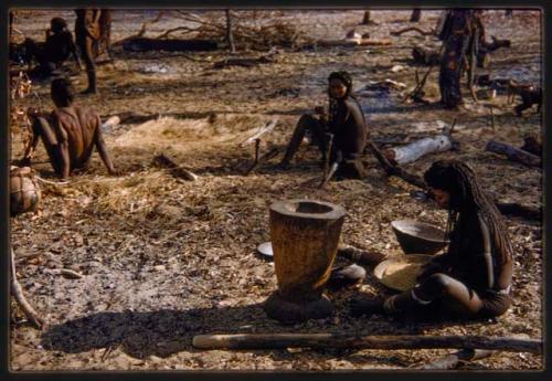 Woman sorting grain, with other people in the background, seated profile