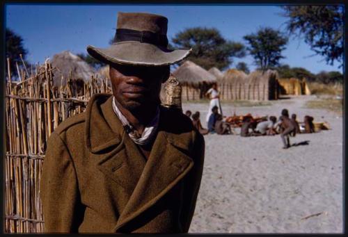 Man standing, with group of people, huts in background