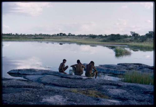 Three boys sitting at the edge of the water, seen from behind