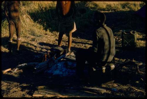 Manuel kneeling by a campfire, with two people standing near him