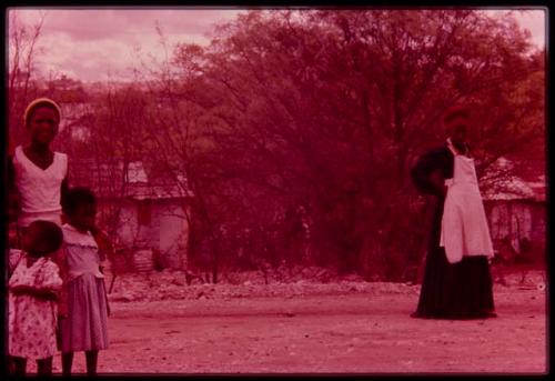 Woman standing in Herero dress, with children in the foreground