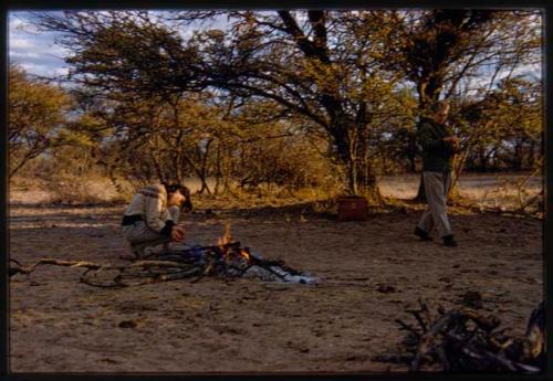 Deborah Marshall squatting near campfire and Lorna Marshall walking by