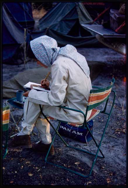 Deborah Marshall in a tsetse fly protective clothing, sitting on a chair and taking notes