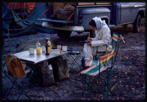 Deborah Marshall in a tsetse fly protective clothing, sitting on a chair and taking notes; camping car and gear shown in the background