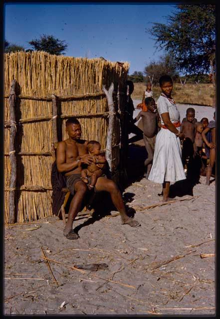 Man sitting in a chair in front of hut and holding baby, with presumably his wife and children near him