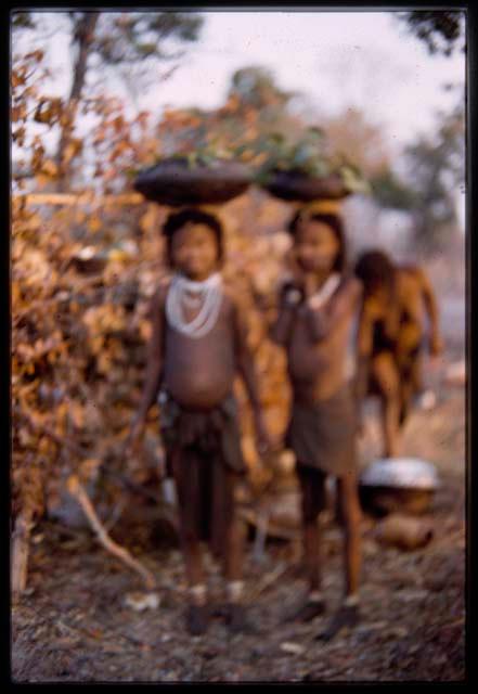 Two girls carrying bowls on their heads (image obscured)