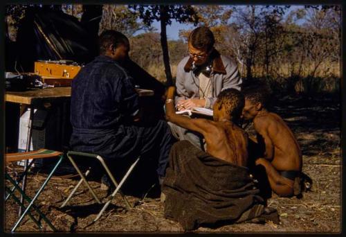 Nicholas England writing in a notebook, sitting with Kernel Ledimo, Hu/Na, and Kumsa