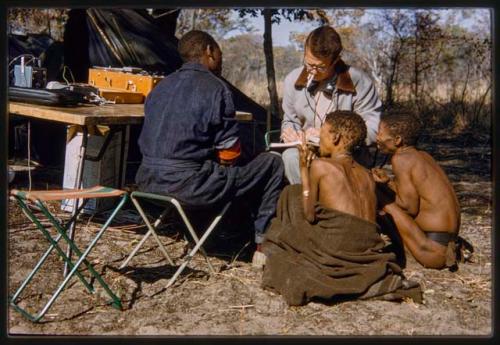 Nicholas England writing in a notebook, sitting with Kernel Ledimo, Hu/Na, and Kumsa