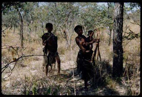 Fiber Root: //Kushay standing, holding fiber roots and carrying a child, with !U standing behind her